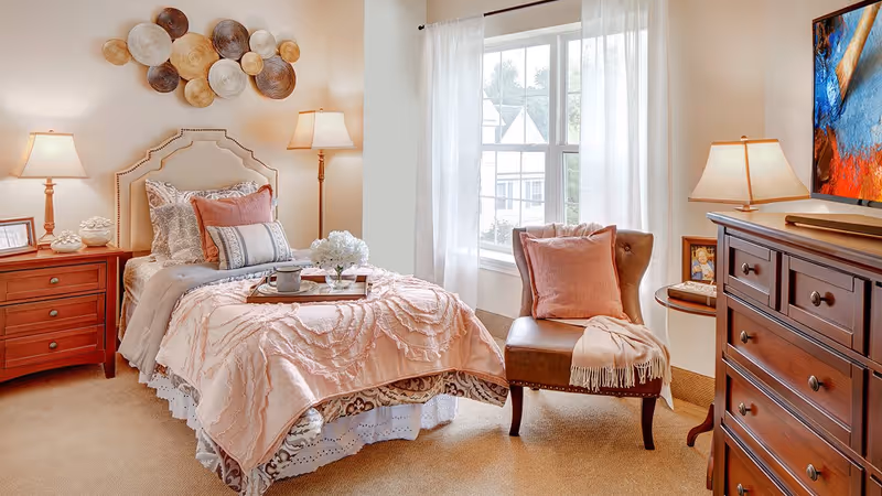 A cozy bedroom with a single bed adorned with multiple pillows and a pink textured blanket. There is a wooden nightstand with a lamp and decorative items on the left side of the bed. A cushioned chair with a pink pillow and a throw blanket is placed near a window with sheer white curtains. On the right side, there is a wooden dresser with a lamp and a colorful abstract painting hanging above it. The room has warm lighting and neutral tones.