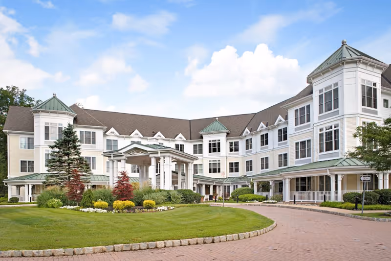 Exterior view of a large, three-story senior living facility building with white siding and green roofs, surrounded by well-maintained landscaping including trees, shrubs, and flowers, under a partly cloudy blue sky.