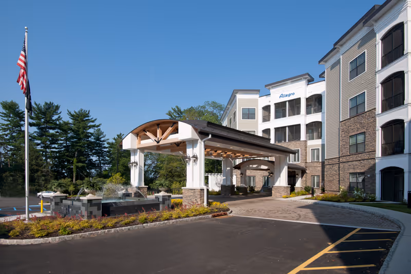 Exterior view of Allegro Senior Living in Harrington Park, NJ, showing a multi-story building with balconies, a covered entrance with wooden beams and stone pillars, a fountain, landscaped garden beds, and an American flag on a flagpole under a clear blue sky.