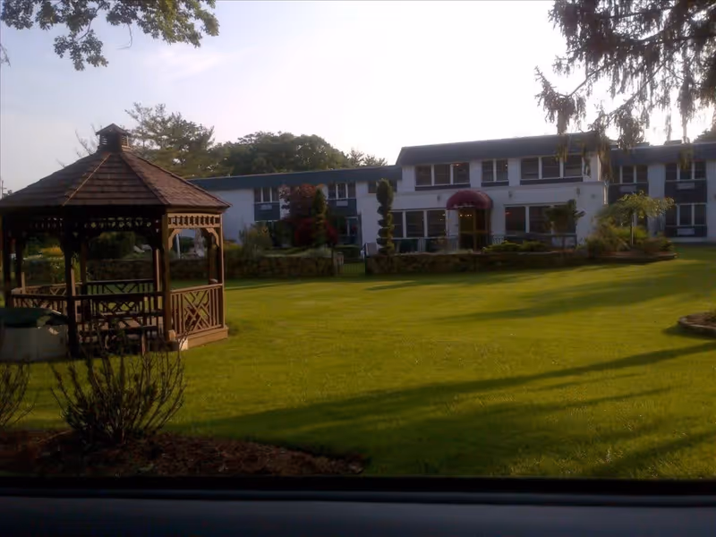 A well-maintained outdoor garden area with a wooden gazebo on the left side and a two-story building in the background. The building has multiple windows and a red awning over the entrance. The lawn is green and neatly trimmed, with some bushes and trees around the garden.