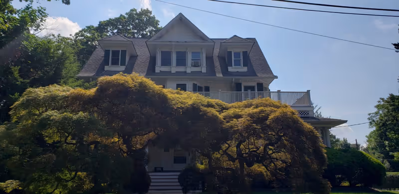 A large, multi-story house with a steep roof and dormer windows partially obscured by a large, leafy tree in the front yard under a clear blue sky.
