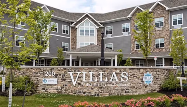 Stone entrance wall reading 'VILLAS' in front of a three-story senior living building with landscaping and flowering shrubs.