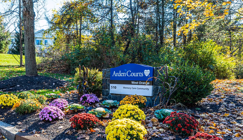 A landscaped garden area with colorful flowers including yellow, red, and purple blooms surrounding a stone sign that reads 'Arden Courts West Orange Memory Care Community 510'. Trees and bushes are visible in the background with a building partially visible behind the trees.
