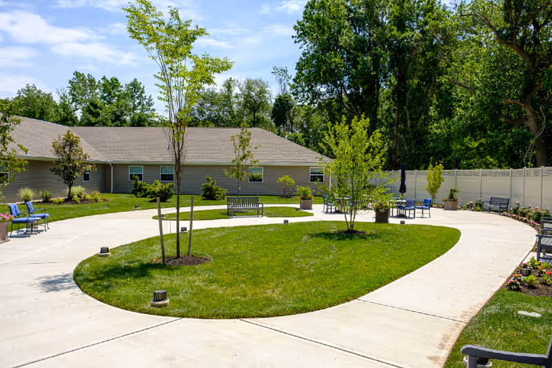 Outdoor courtyard area at a senior living facility with a curved concrete walkway surrounding a grassy area with small trees and benches. The background shows a single-story building and a white privacy fence with trees beyond it under a blue sky with some clouds.