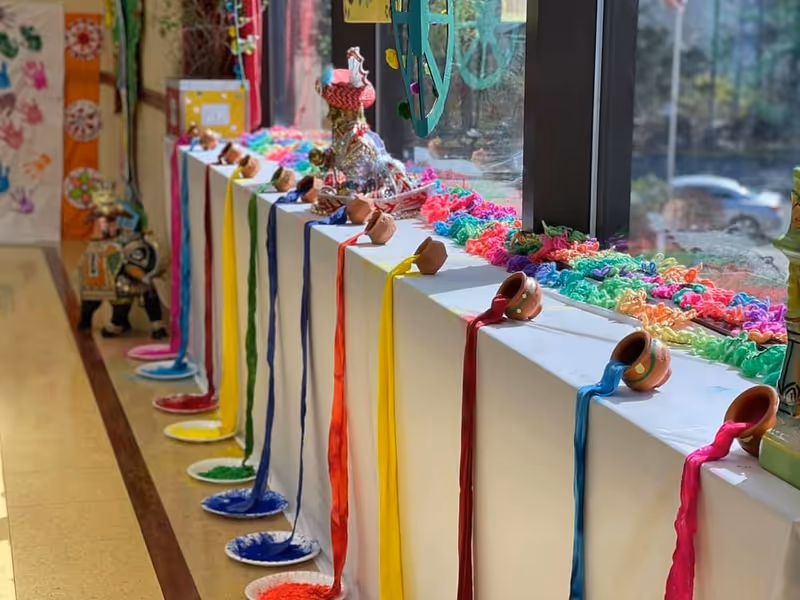 A colorful indoor display along a windowsill featuring small clay pots tilted to pour vibrant colored powders onto white plates below. The powders are arranged in a rainbow sequence, with additional colorful decorations and figurines placed along the windowsill.