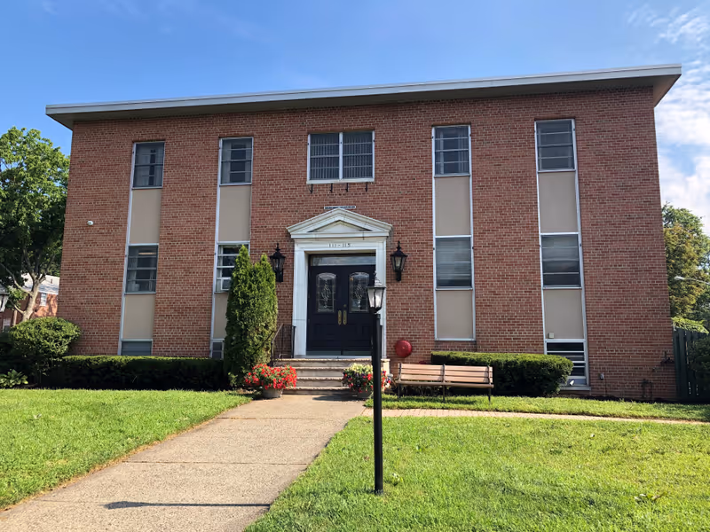 Front exterior of a two-story brick building with a central doorway, walkway, lawn, and bench.