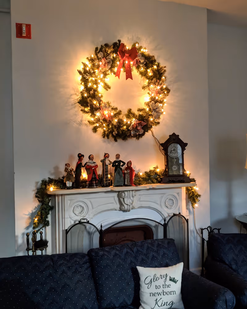 A cozy living room area with a dark blue couch featuring a pillow that says 'Glory to the newborn King.' Above the white ornate fireplace mantel is a large Christmas wreath decorated with lights and a red bow. The mantel is adorned with small figurines and a vintage clock, with additional greenery and lights draped around it.