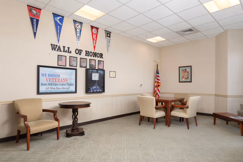 A room with beige walls and carpet featuring a 'Wall of Honor' display dedicated to veterans, including framed photos, flags, and military pennants. The room has several beige upholstered chairs, a round wooden table, a rectangular wooden table with four chairs, and an American flag in the corner.