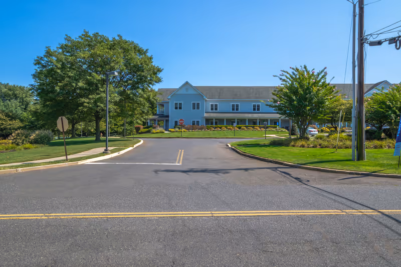 Exterior view of a two-story light blue building with a porch, surrounded by green trees and landscaping under a clear blue sky. The building is set back from a paved road with a double yellow line and a stop sign visible near the entrance driveway.