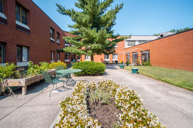 Outdoor courtyard area at a senior living facility with brick buildings surrounding a paved walkway. There are green metal chairs and a table, raised garden beds with plants, bushes, and a large tree in the center. The sky is clear and blue.