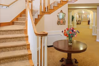 Interior view of a senior living facility hallway with carpeted stairs on the left, a round wooden table with a vase of colorful flowers in the center, and a corridor leading to seating areas and rooms in the background.