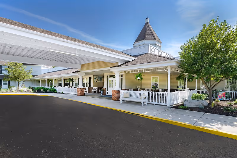 Exterior view of Mattison Crossing facility showing a covered entrance with white columns and a porch area with white benches and chairs. The building has beige siding, a peaked roof with a small tower, and is surrounded by trees and landscaping under a clear blue sky.