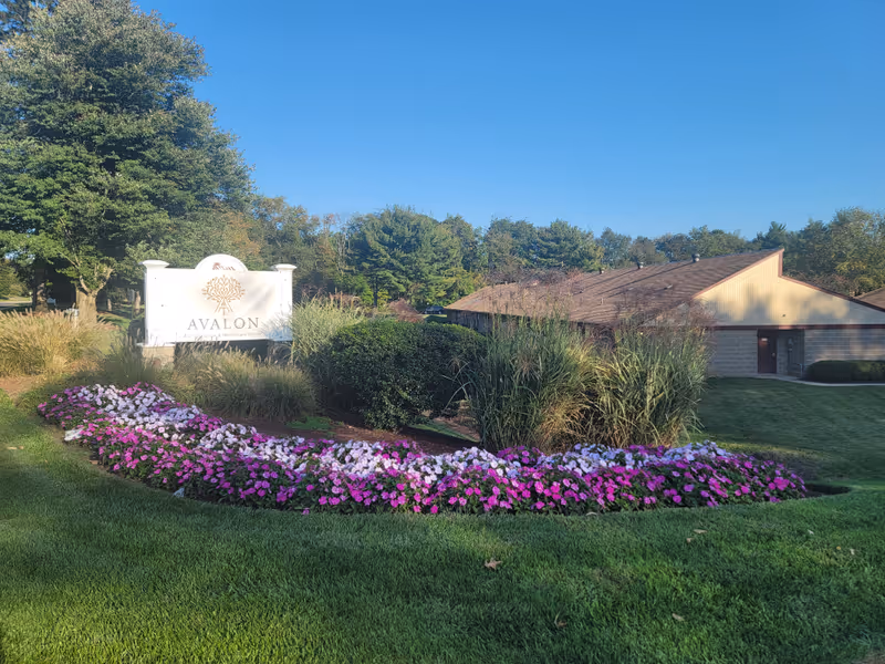 Outdoor view of Avalon Rehabilitation & Healthcare Center featuring a landscaped garden with pink and white flowers, green bushes, and tall grasses in front of a building with a sloped roof under a clear blue sky.