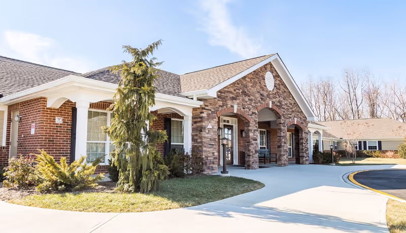 Exterior view of a senior living facility building with a brick and stone facade, a covered entrance with arches, a sidewalk, and some landscaping including bushes and a tree. The sky is clear and blue.