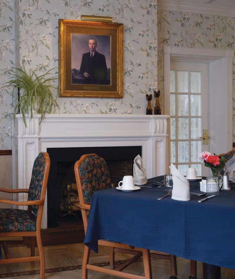 Dining room with a set table and upholstered chairs in front of a white fireplace topped by a framed portrait and floral wallpaper.