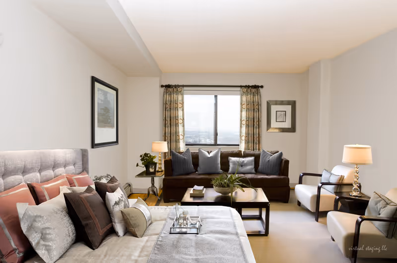 A well-lit living room with a large window in the background. The room features a brown sofa with multiple gray and blue pillows, two white armchairs with dark wooden arms, a coffee table with a plant and books, and a bed with various pillows in the foreground. There are two table lamps on either side of the sofa and armchairs, framed artwork on the walls, and patterned curtains on the window.