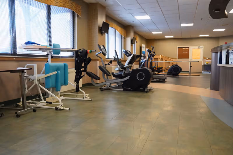 A bright rehab and exercise room with stationary bikes, therapy equipment, and large windows.