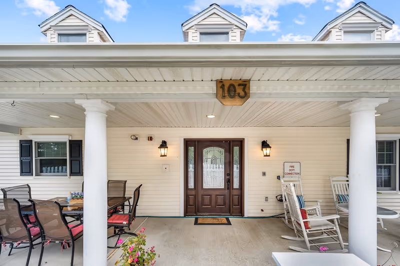 Front porch of a building with white siding and a dark wooden door with decorative glass panels. The porch has white columns, outdoor seating including a table with chairs and rocking chairs, and a wooden sign above the door displaying the number 103. There are two wall-mounted lantern-style lights on either side of the door and a fire department connection sign on the right wall.