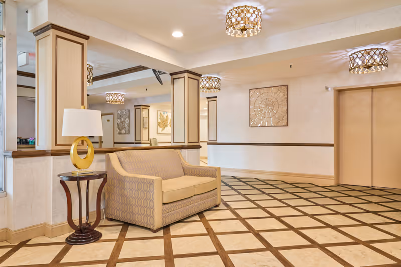 A well-lit interior common area with a patterned tile floor, beige walls, and decorative ceiling lights. There is a beige patterned loveseat next to a small round wooden table with a modern lamp on it. The area includes mirrored columns and framed artwork on the walls. Two closed elevator doors are visible on the right side.