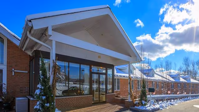 Exterior view of Bentley Assisted Living at Branchville on a sunny winter day with snow on the ground and roof. The building is made of red brick with large windows and a covered entrance. The sky is blue with some clouds.