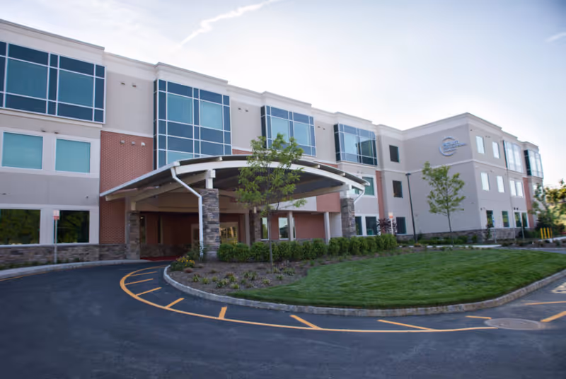 Exterior view of a modern three-story building with large windows and a covered entrance. The building is surrounded by a well-maintained lawn, small trees, and shrubs. The driveway curves around the entrance with marked parking spaces.