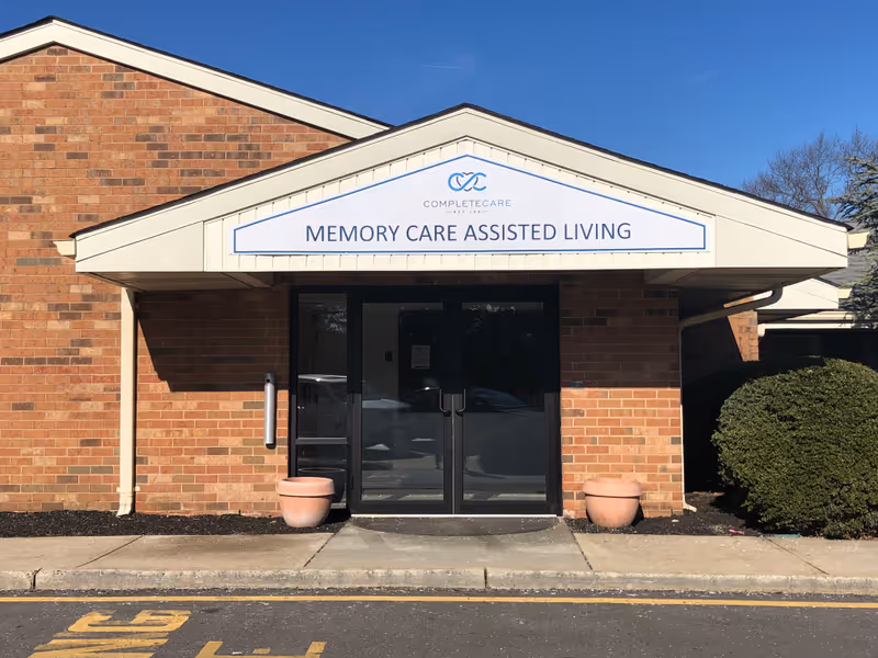 Front entrance of a brick building with a white triangular awning displaying the sign 'Complete Care at Bey Lea MEMORY CARE ASSISTED LIVING'. There are two large glass doors, two empty flower pots on either side of the entrance, and a bush to the right. The sky is clear and blue.
