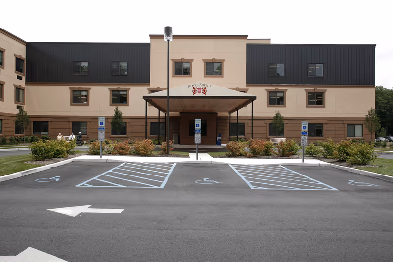 Front exterior view of Royal Suites Healthcare & Rehabilitation Center showing the building entrance with a covered drop-off area, several windows, landscaped bushes, and three handicap parking spaces in front.