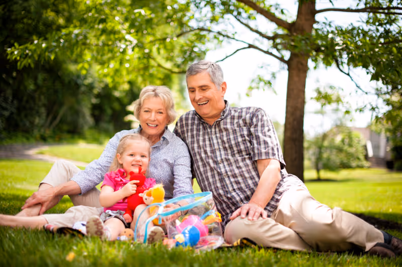 An elderly couple sitting on the grass outdoors with a young girl, smiling and enjoying a sunny day surrounded by trees and greenery. The girl is holding colorful toys.