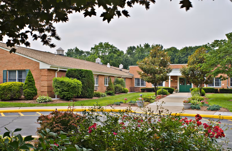 Exterior view of Ridgewood Center, a single-story brick building with green doors and windows, surrounded by well-maintained landscaping including bushes, trees, and flowering plants. A paved walkway leads to the entrance, and the sky is overcast.