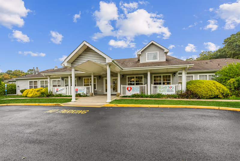 Front exterior view of The Addison of Somers Place building with a covered entrance, white columns, and banners expressing appreciation for staff. The building is surrounded by green bushes and a clear blue sky with some clouds.