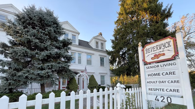 Exterior view of a white multi-story senior living facility named Beverwyck House with a white picket fence and large evergreen trees in front. A sign near the entrance displays services including Senior Living, Home Care, Adult Day Care, and Respite Care. The sky is clear and blue.