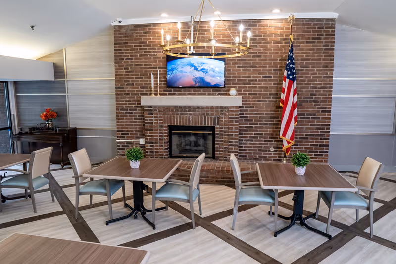 A cozy common area with a brick fireplace, a mounted flat-screen TV above it displaying an image of Earth, and an American flag standing to the right. The room has several square tables with chairs around them, each table decorated with a small green plant. The floor has a light wood pattern with darker wood accents, and there is a chandelier hanging from the ceiling.