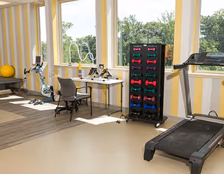 Bright exercise/therapy room with a treadmill, stationary bike, dumbbell rack, chairs and large windows overlooking trees.