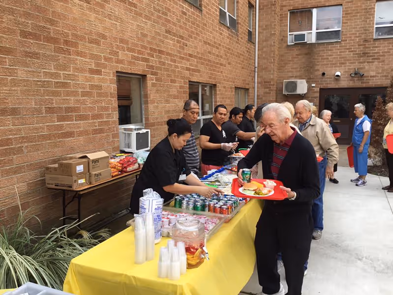 A group of elderly people and staff members are gathered outdoors at Lakeland Health Care Center. They are serving and receiving food and drinks from a long table covered with a yellow tablecloth. The table has various canned sodas, cups, and food items. The setting is in a courtyard area surrounded by brick walls of the building.