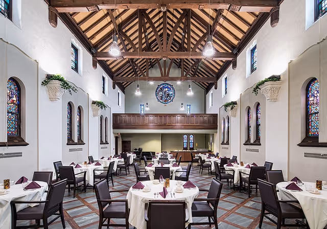 Large dining room with round tables set with white linens and purple napkins inside a high‑ceilinged, chapel‑like hall with stained glass windows and exposed wooden beams.