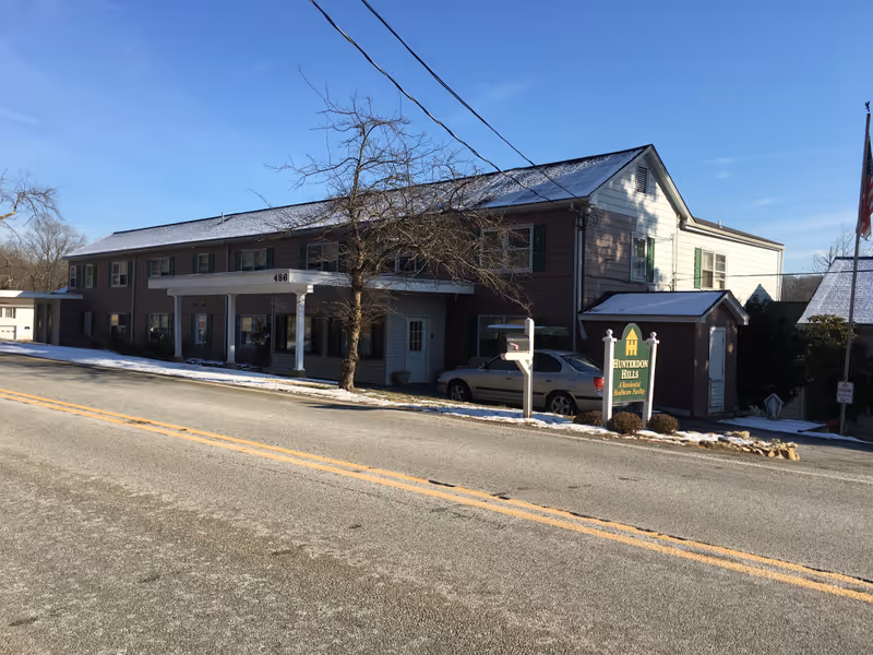 Exterior view of Hunterdon Hills Residential Care Facility, a two-story building with a covered entrance, a tree without leaves in front, a parked car, and a sign displaying the facility's name. The ground and roof have a light dusting of snow, and the sky is clear and blue.