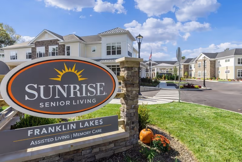 Exterior view of Sunrise Senior Living facility in Franklin Lakes with a large sign in the foreground displaying the facility name and services. The building is two stories with white and stone facade, surrounded by a well-maintained lawn and a clear blue sky with some clouds.