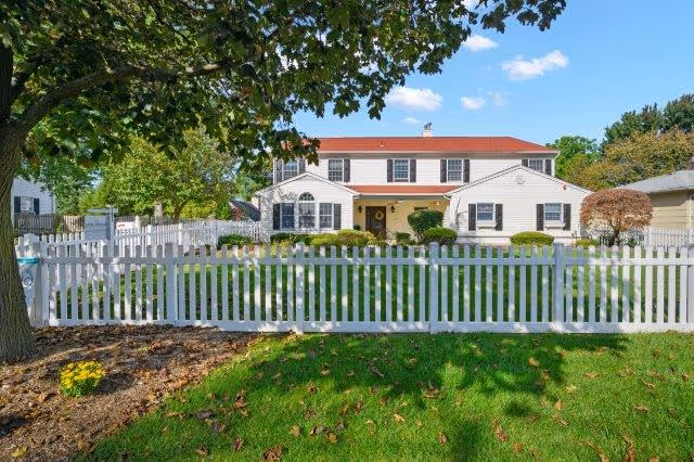 A two-story white house with a red roof behind a white picket fence. The house is surrounded by a well-maintained lawn, bushes, and trees under a blue sky with some clouds.
