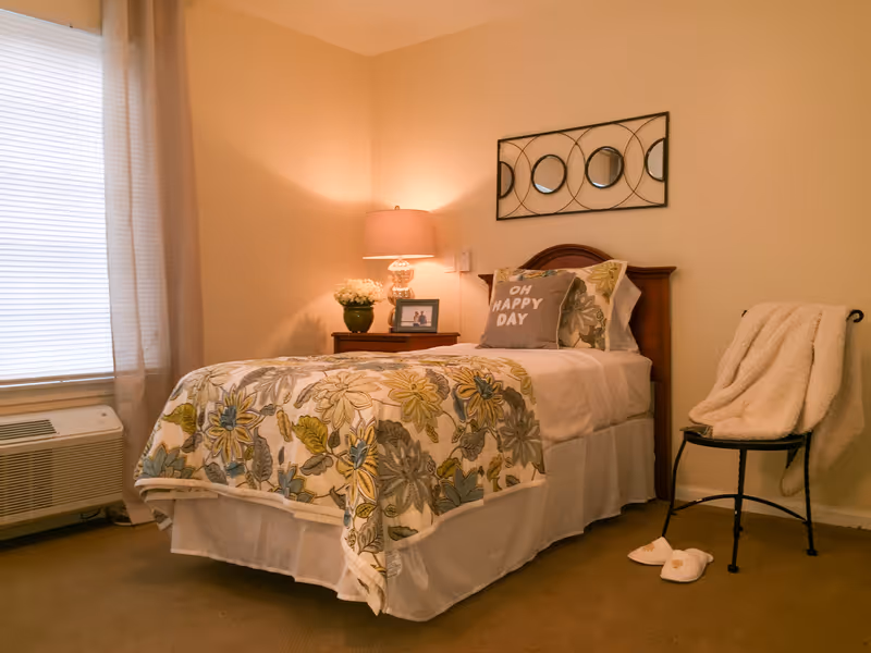 A cozy bedroom with a single bed covered in a floral patterned quilt and pillows, including one with the text 'OH HAPPY DAY'. Next to the bed is a wooden nightstand with a lamp, a small green plant, and a framed photo. A decorative wall mirror with circular designs hangs above the bed. A chair with a soft blanket draped over it and a pair of slippers on the floor are also visible. The room has beige walls, a window with blinds and sheer curtains, and a carpeted floor.