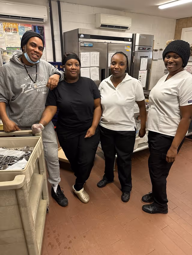 Four staff members standing together in a kitchen area. They are dressed in casual and work attire, with kitchen equipment and stainless steel refrigerators in the background.