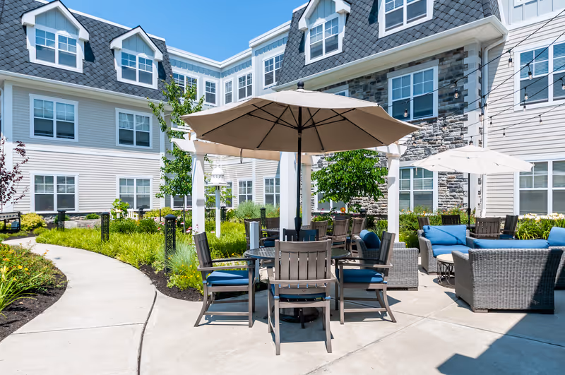 Outdoor patio area at Arbor Terrace Mountainside featuring a round table with wooden chairs and blue cushions under a large beige umbrella. Surrounding the patio are additional seating areas with wicker furniture and blue cushions. The patio is bordered by a curved concrete walkway and landscaped greenery. The background shows a multi-story building with gray siding, stone accents, and multiple windows under a clear blue sky.