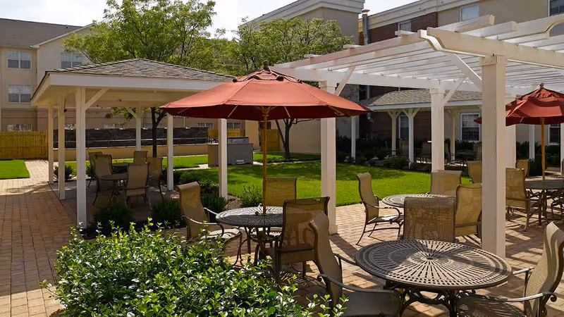 Outdoor patio area at Atria Cranford featuring multiple round metal tables with mesh chairs and red umbrellas. The space includes white pergolas and a covered seating area with additional tables and chairs. Surrounding the patio are green lawns, shrubs, and trees, with parts of the building visible in the background.