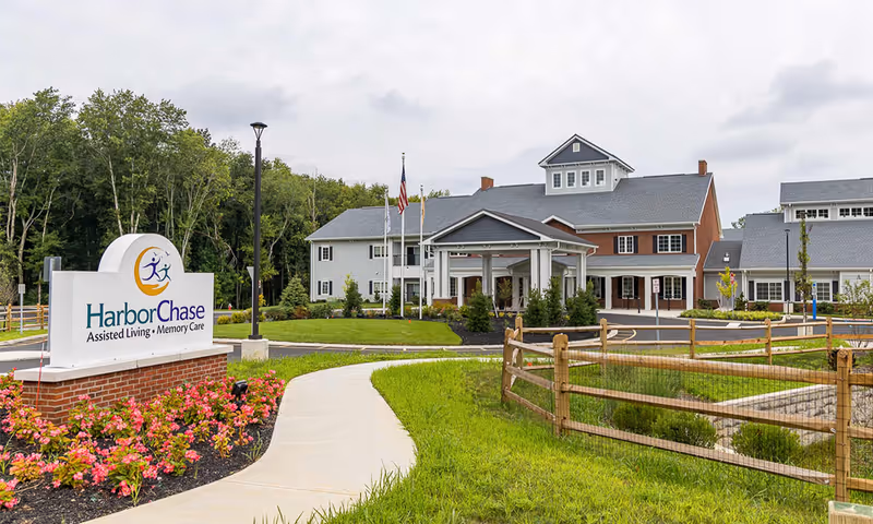 Exterior view of HarborChase Of Princeton assisted living and memory care facility with a paved walkway, landscaped garden with flowers, a wooden fence, and a large building with a covered entrance and multiple windows under a cloudy sky.