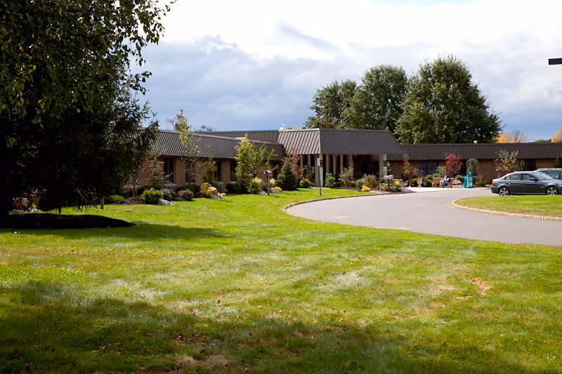 Single-story care facility building with a landscaped lawn, trees, and a curved driveway with a parked car.