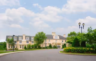 Front view of a three-story beige health center with a circular driveway, landscaped island and lamppost under a cloudy sky.