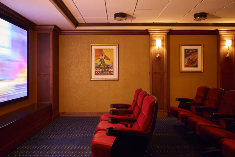 Small private theater room with rows of red upholstered seats facing a large screen, wood-paneled walls, framed posters, and wall sconces.