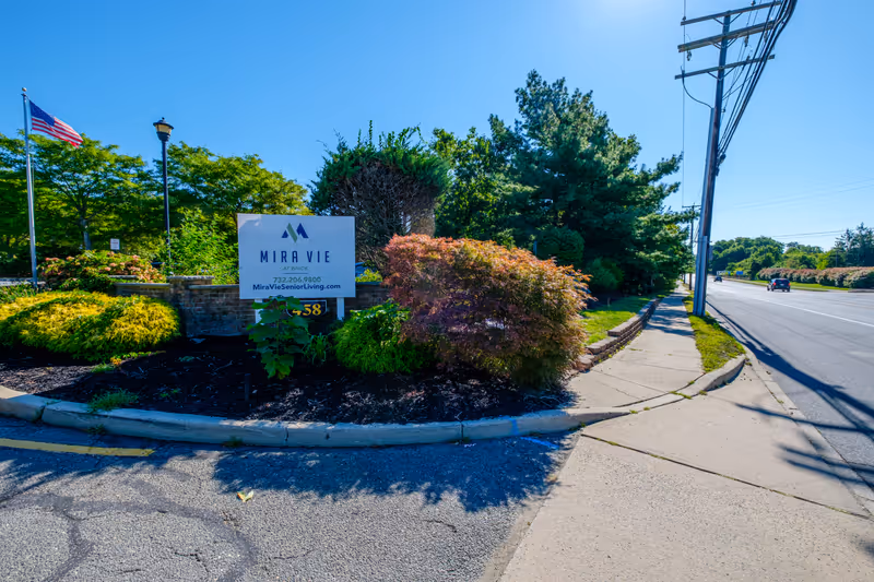 Entrance sign reading 'Mira Vie' amid landscaped shrubs next to a sidewalk and road with an American flag in the background.