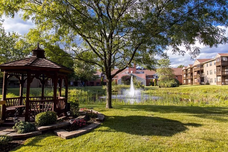 A peaceful outdoor scene at The Fountains at Atco featuring a wooden gazebo surrounded by greenery, a large tree, a pond with a water fountain, and residential buildings in the background under a partly cloudy sky.