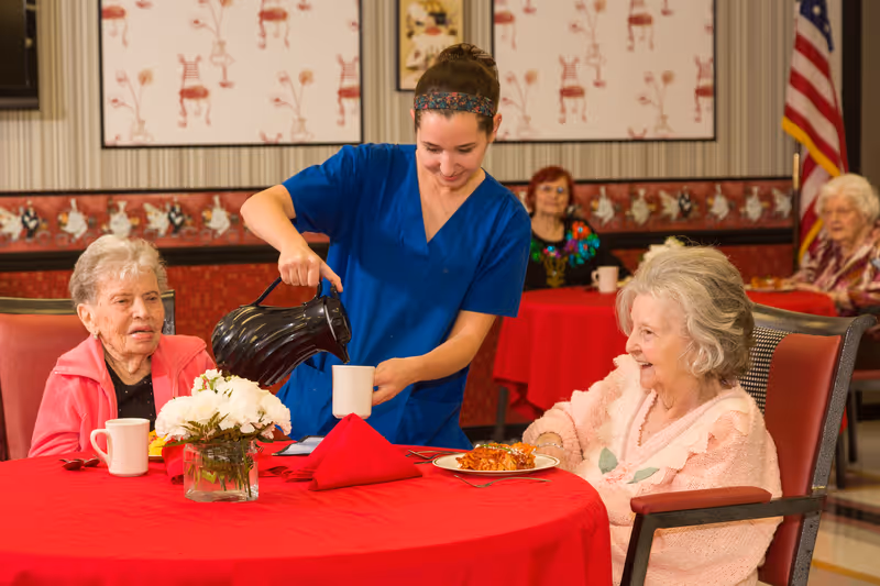 A young caregiver in blue scrubs is pouring coffee into a mug for an elderly woman seated at a table with a red tablecloth. Another elderly woman sits nearby, and two more elderly women are visible in the background, all in a dining room setting with patterned wallpaper and an American flag in the corner.