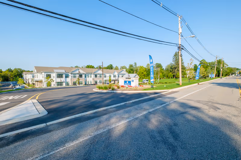 Exterior view of Arbor Terrace Basking Ridge senior living facility on a clear sunny day, showing a two-story building with a parking lot to the left and a landscaped area with flags that say 'NOW OPEN' and 'TOURS AVAILABLE' near the entrance.